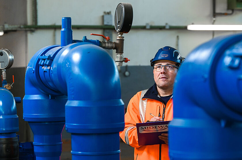 Wasserwerk Pratau Ein MIDEWA-Mitarbeiter mit orangener Arbeitskleidung steht in einem Technikraum eines Wasserwerkes. Er befindet sich auf einem Kontrollgang und notiert technische Werte auf einem Formular. Er trägt eine Brille und einen blauen Schutzhelm.