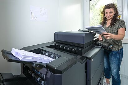 Druckzentrum Eine Mitarbeiterin in blauer Jeans und gemusterter Bluse hält einen Papierstapel in der Hand und steht vor einem großen grauen Bürodrucker. Ein dicker Papierstapel ist im Ablagefach des Druckers zu sehen.