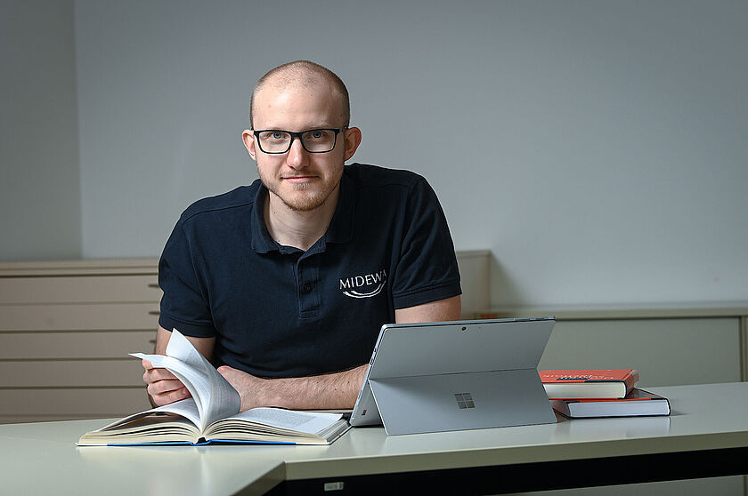 Student Ein junger Mann im blauen T- Shirt sitzt am Schreibtisch und blättert in einem Fachbuch.Er trägt eine schwarze Brille. Neben ihm stehen ein Tablet und zwei weitere Fachbücher mit roter Bucheinbindung.