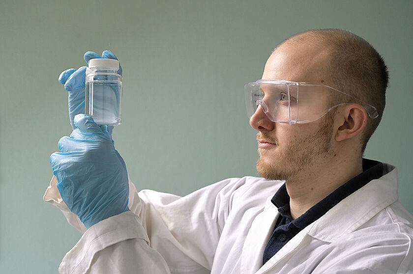 Trinkwasseranalytik Ein Mitarbeiter im weißen Kittel und transparenter Schutzbrille, hält eine Wasserprobe aufbewahrt in einem weißen Glas, in Augenhöhe. Im Rahmen einer Wasseranalyse prüft er Trübung und Färbung der Wasserprobe.