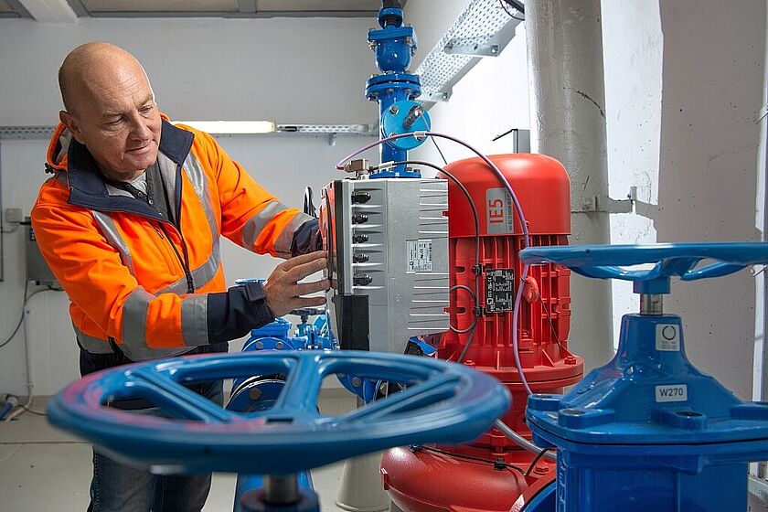 Ein Tinkwassermonteur in orangener Arbeitsjacke, prüft bei einem Kontrollgang in einem Pumpenwerk die technischen Anlagen. Im Vordergrund ist Pumpentechnik in blau und rot zu sehen.