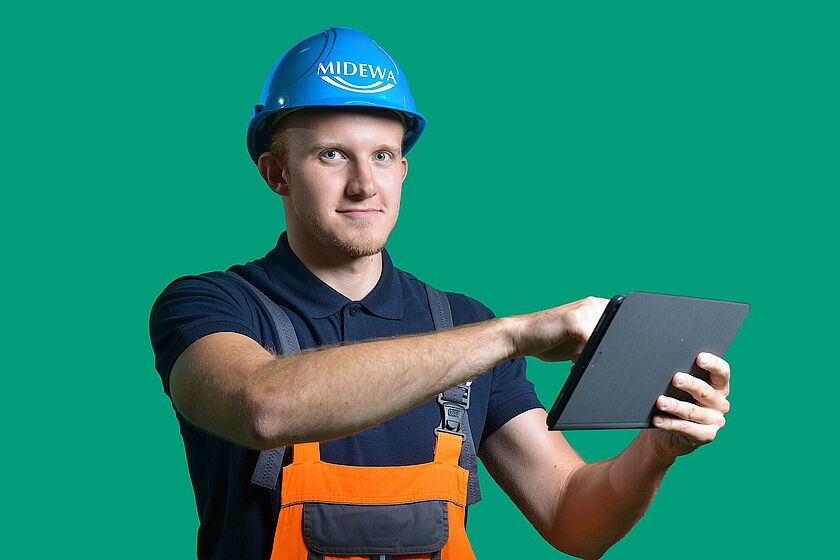 Ein junger Mann in orangefarbener Latzhose und blauem Schutzhelm präsentiert aufmerksam Informationen auf seinem Tablet.