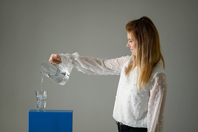 Kundenservice Eine Kundenservicemitarbeiterin schenkt mit einer Glaskaraffe Trinkwasser in ein Trinkglas. Das Trinkwasserglas ist graviert mit dem Logo des Trinkwasserversorgers MIDEWA.