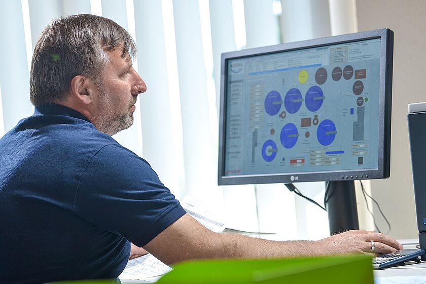 Kläranlage Rollsdorf Ein Mann im blauen T-Shirt sitzt an einem Computer und schaut auf eine eingeblendete Grafik aus dem Abwasserbereich.